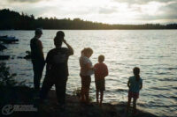 Colour film photo of a lake with trees on the horizon. In the foreground are five silhouettes looking out on the water, they're mostly in shadow.