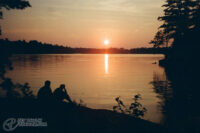 Colour film photo of a very orange sunset over a lake, there's two dark silhouettes in the bottom looking off at the sunset.