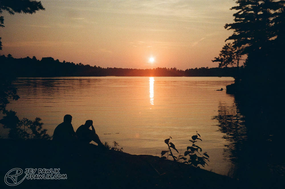 Colour film photo of a very orange sunset over a lake, there's two dark silhouettes in the bottom looking off at the sunset.